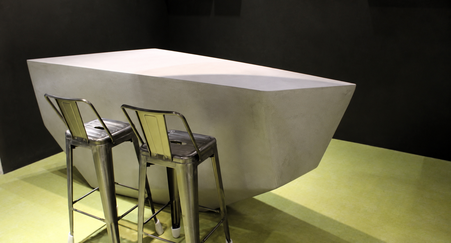 Mesa-isla facetada de microcemento gris con taburetes metálicos sobre suelo verde, portada de proyectos TECNOCEMENTO